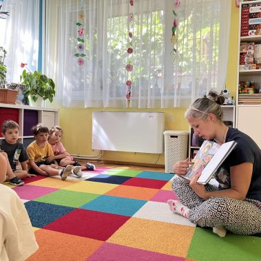 Авторката на книгата показва илюстрация от книгат ана деца от детска градина седнали в кръг около нея