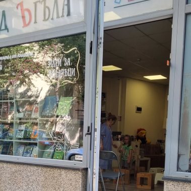 Вход на книжарница в който се виждат подредено пространство за четене с деца