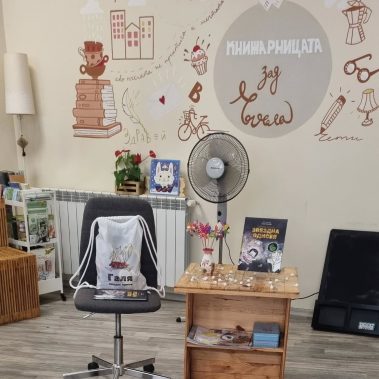 Книгата Звездна одисея на маса с лампички, до нея раница с илюстрация на коте снимани на фона на интериора на книжарница зад ъгъла