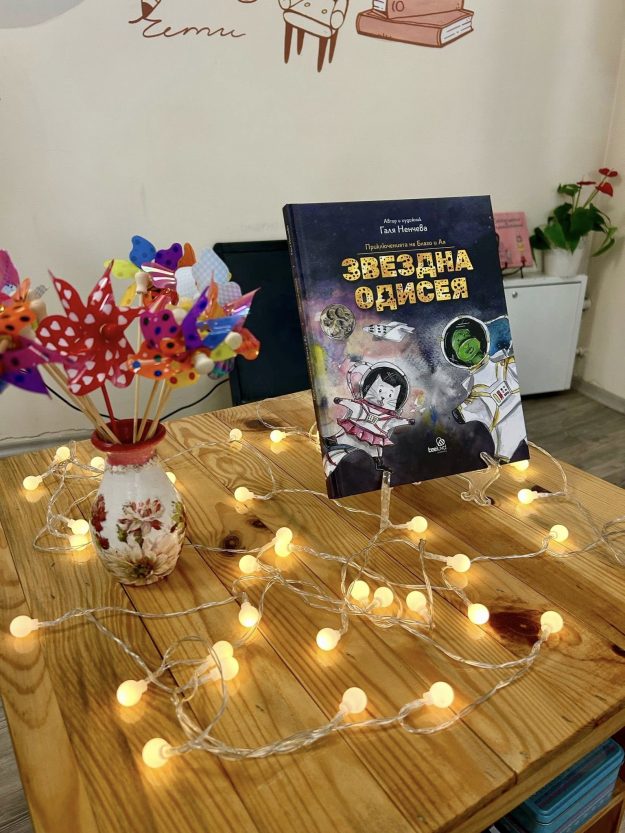 Книгата звездна одисея в книжарницата детската книга за Космоса Звездна одисея изложена на маса с малки лампички около нея