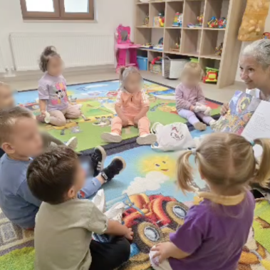 Деца, седнали в кръг около жена, която им показва ярки илюстрации в книга за Космоса