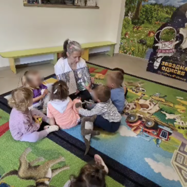 Деца са се скупчили около жена, която държи книга и показват на илюстрацията къде има коте