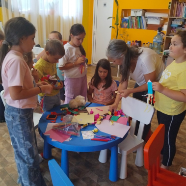 Художничката Галя Ненчева наведена над маса с материали за изработване на ракета от хартия, заобиколена от децата на които помага