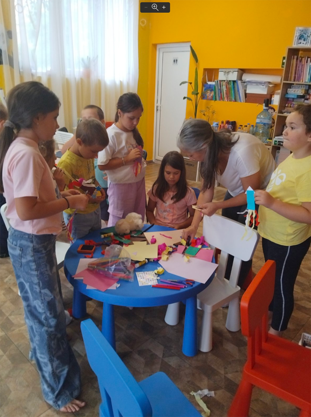 Художничката Галя Ненчева наведена над маса с материали за изработване на ракета от хартия, заобиколена от децата на които помага