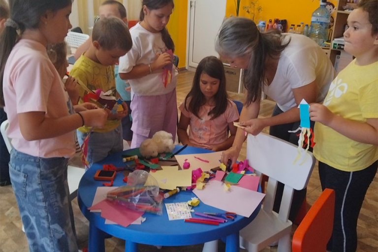 Художничката Галя Ненчева наведена над маса с материали за изработване на ракета от хартия, заобиколена от децата на които помага