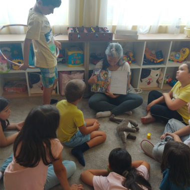 Авторката на Звездна одисея заобиколена от насядали по земяат деца, които гледат илюстрацията, която тя им показва